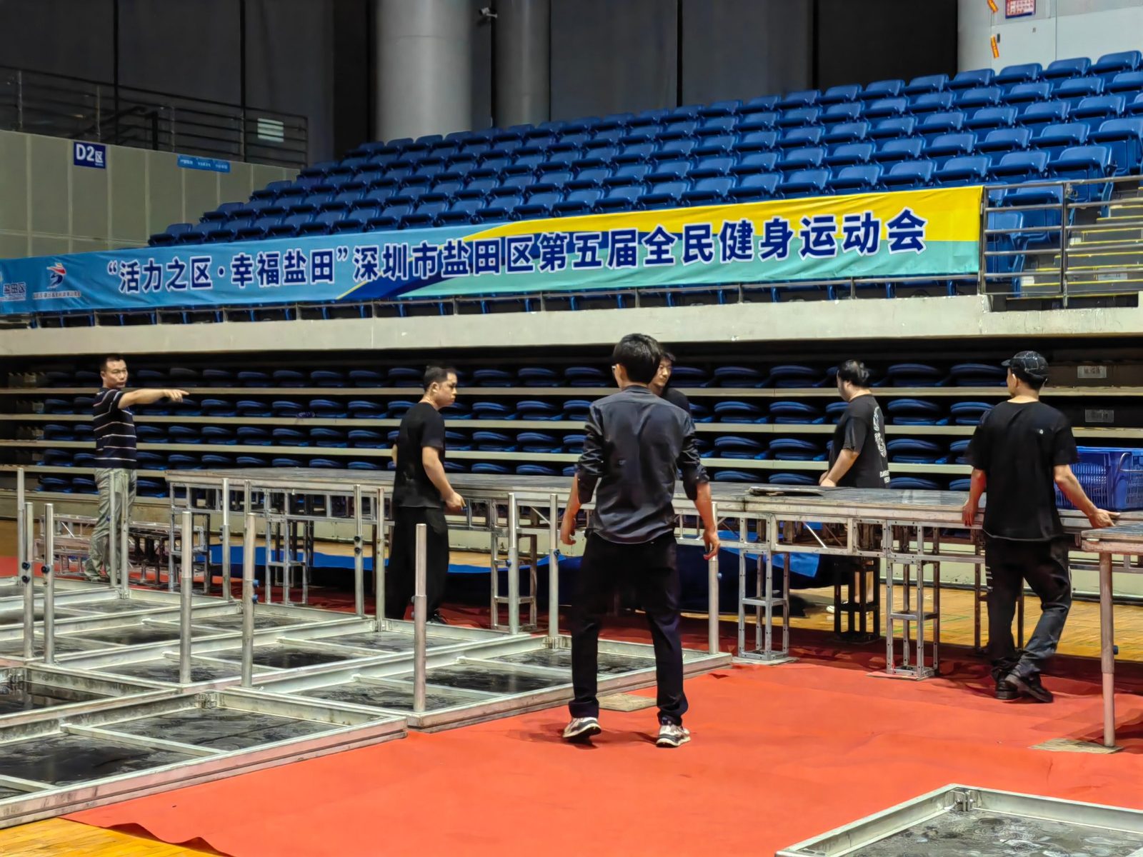 深圳体育馆舞台搭建现场：工人在看台下方组装铝合金桁架模块，红毯地面与观众席